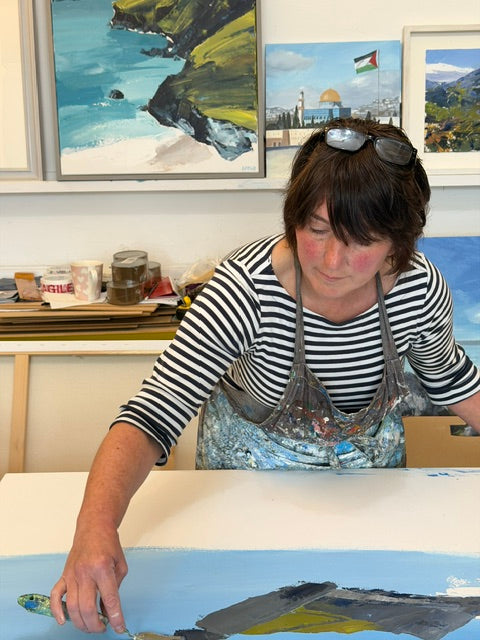 A woman wearing a black and white striped shirt and a paint-splattered apron paints a landscape painting on a white canvas in an art studio. Framed coastal and architectural paintings hang on the wall behind her.