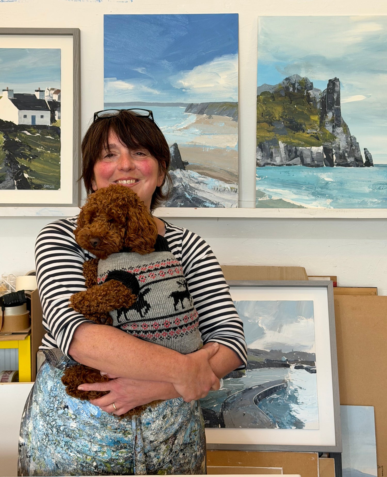 A smiling woman with short brown hair stands indoors holding a small brown poodle. She wears a black-and-white striped long-sleeve top and a gray patterned sweater with black dog silhouettes.